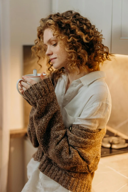 a woman standing in a kitchen holding a cup of coffee, by Adam Marczyński, trending on pexels, renaissance, brown curly hair, brown sweater, ethereal soft and fuzzy glow, thinking pose