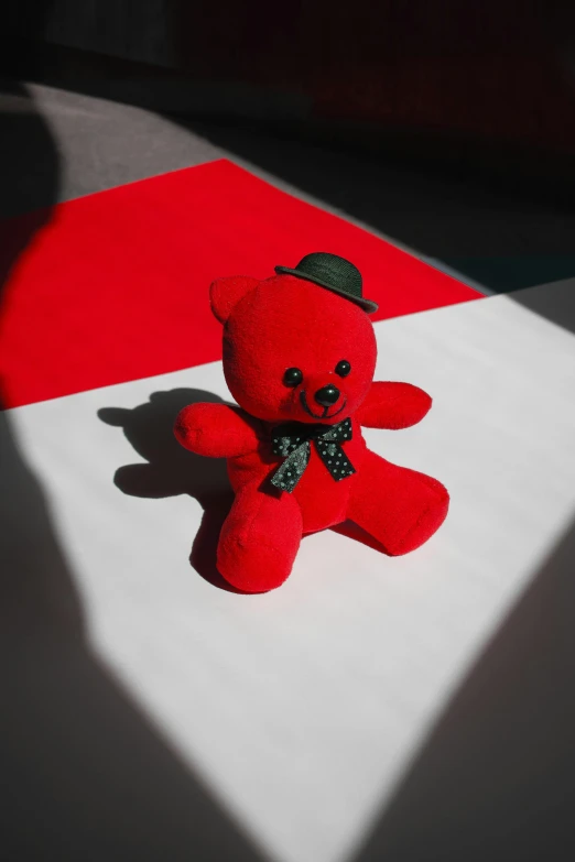 a red teddy bear sitting on top of a table, in red and black, sitting on the floor, in the sun