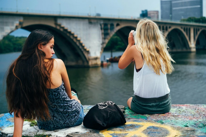 a couple of women sitting on top of a cement wall, a photo, trending on pexels, happening, bokeh on side of the river, college girls, big arches in the back, a blond