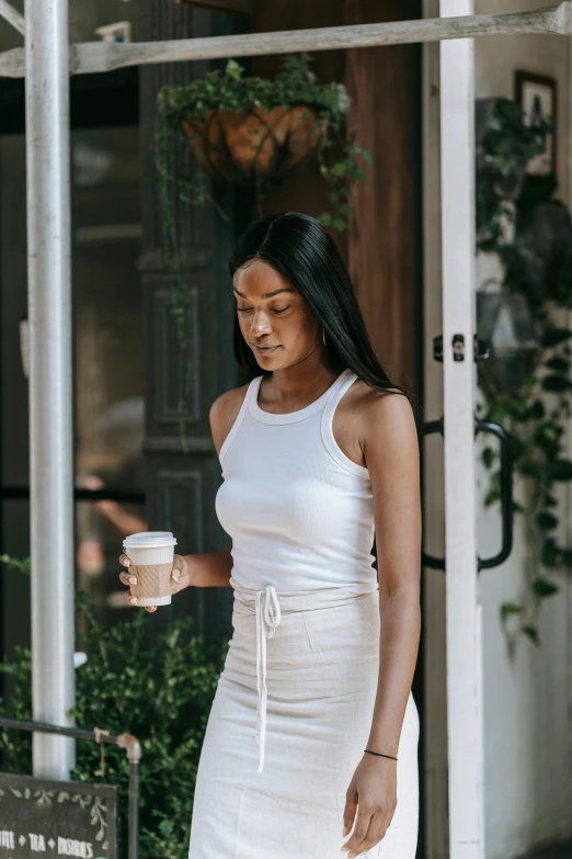 a woman in a white dress holding a cup of coffee, by Nina Hamnett, trending on unsplash, wearing a tanktop and skirt, tan skin a tee shirt and shorts, walking confidently, off - white collection