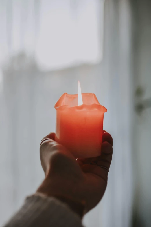 a person holding a candle in front of a window, inspired by Elsa Bleda, pexels, pale red, made of glazed, on a gray background, orange hue