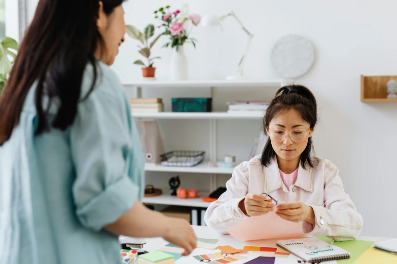 a woman sitting at a table looking at a piece of paper, a cartoon, pexels contest winner, asian woman made from origami, woman holding another woman, studious, gif