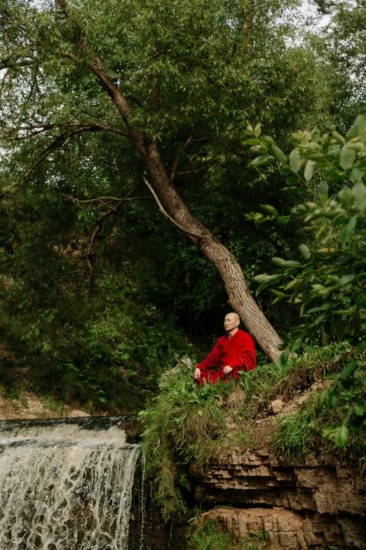 a man sitting on top of a cliff next to a waterfall, an album cover, unsplash, renaissance, wearing red robes, sitting under a tree, marina abramovic, portrait tilda swinton