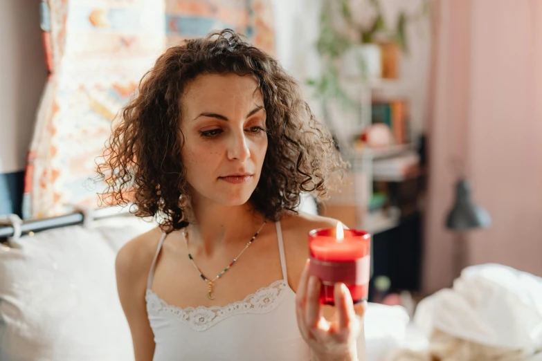 a woman sitting on a bed holding a cup, by Julia Pishtar, trending on pexels, renaissance, holding a candle holder, red aura, worried, curly haired