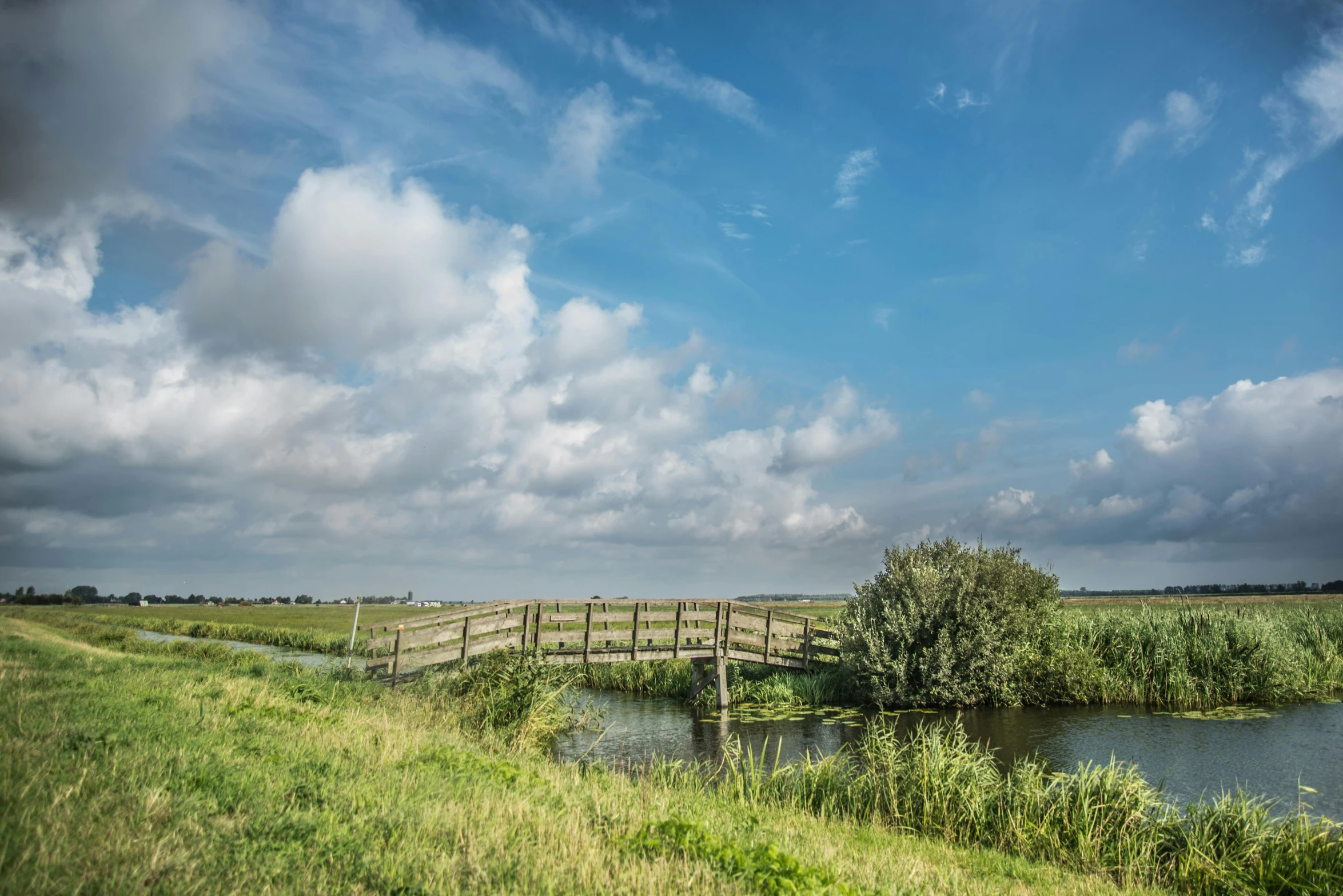 a wooden bridge over a body of water, by Jacob Esselens, unsplash, land art, blue sky and green grassland, dutch masters, thumbnail, canal