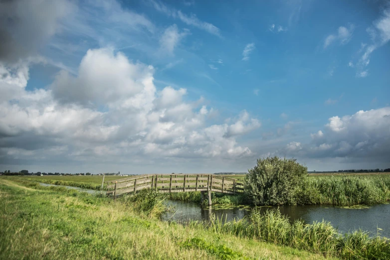 a wooden bridge over a body of water, by Jacob Esselens, unsplash, land art, blue sky and green grassland, dutch masters, thumbnail, canal