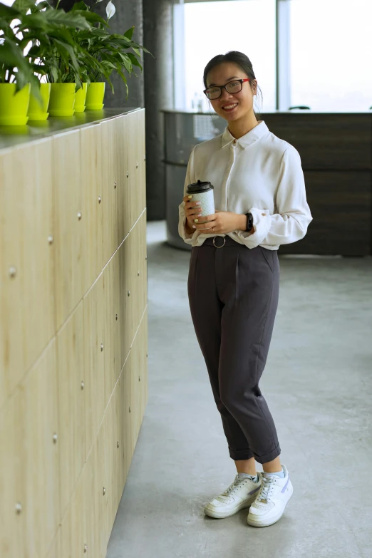 a woman standing next to a counter holding a cup of coffee, inspired by Cheng Jiasui, happening, wearing pants, corporate style, malaysian, at college