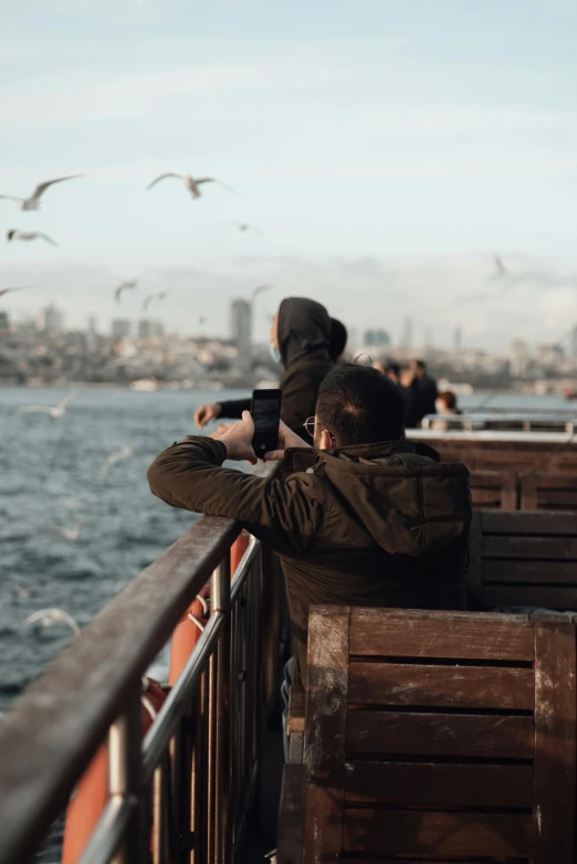 a man sitting on top of a wooden bench next to a body of water, by irakli nadar, pexels contest winner, helicopters and tremendous birds, on ship, people watching around, movie footage