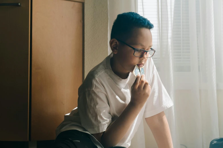 a boy sitting on the floor brushing his teeth, inspired by Liam Wong, pexels contest winner, blue rimmed glasses, window light, profile image, having a snack