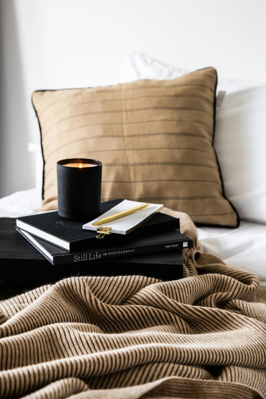 a stack of books sitting on top of a bed, a still life, inspired by Pierre Soulages, reddit, candle, manuka, all black matte product, caramel