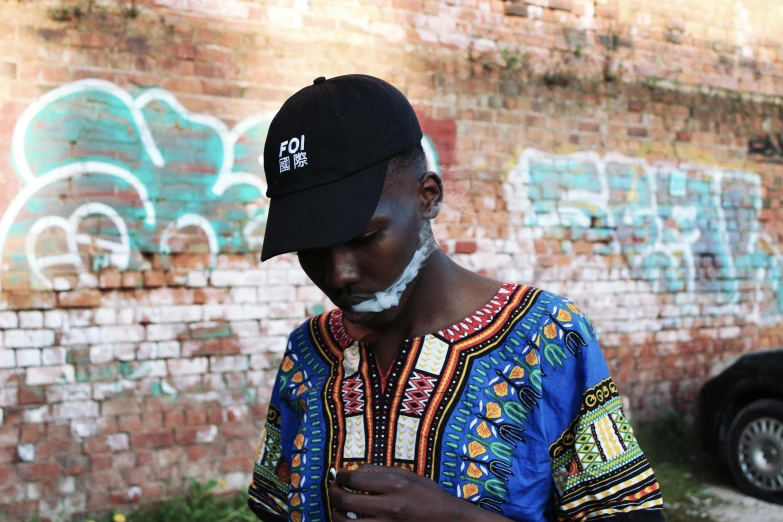 a man standing in front of a brick wall, inspired by Gerard Sekoto, pexels contest winner, afrofuturism, wearing a baseball cap, adut akech, wearing ornate clothing, chewing tobacco