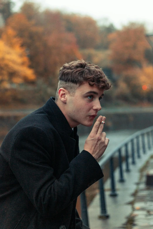 a man standing next to a body of water, an album cover, by Jacob Toorenvliet, pexels contest winner, lips on cigarette, autumn, undercut hairstyle, scotland