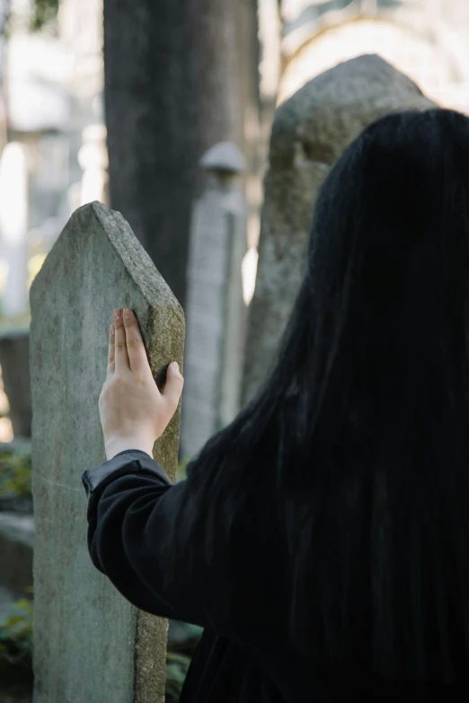 a woman looking at a tombstone in a cemetery, by Attila Meszlenyi, unsplash, with his long black hair, pointing, ignant, an asian woman