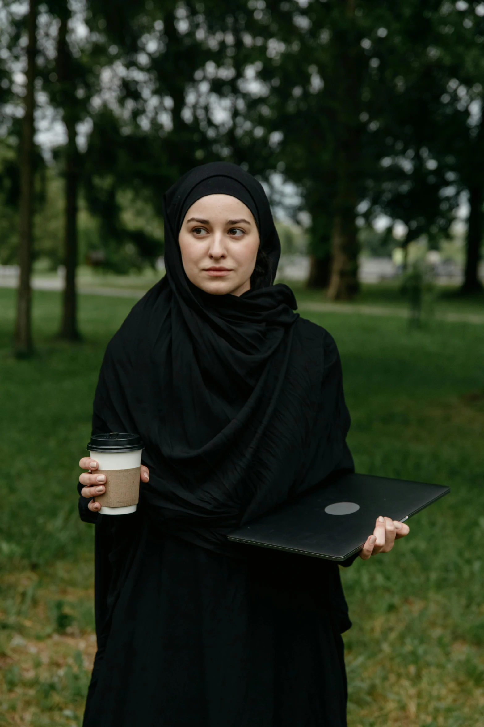 a woman in a black hijab holding a cup of coffee and a laptop, inspired by Modest Urgell, renaissance, 2019 trending photo, drones, a park, ( ( theatrical ) )