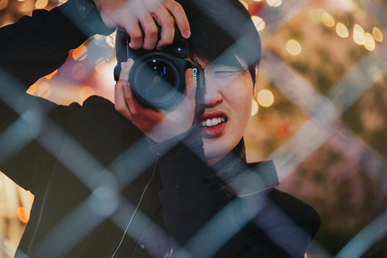 a man holding a camera behind a chain link fence, by Julia Pishtar, pexels contest winner, korean woman, lens zooming, cover photo portrait of du juan, unsplash 4k