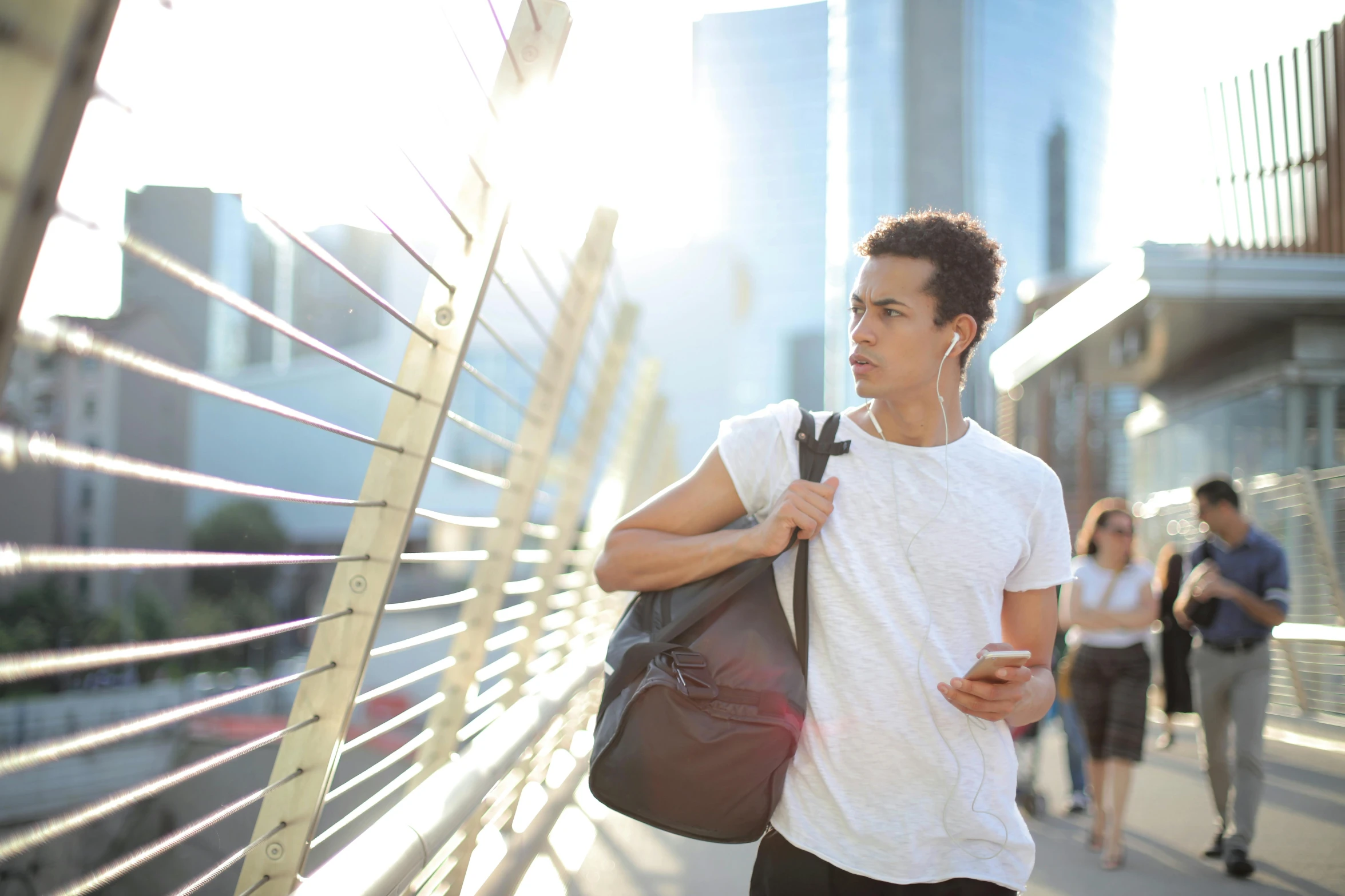 a man with a backpack walking across a bridge, trending on pexels, happening, lean man with light tan skin, looking at his phone, city in the background, aboriginal australian hipster