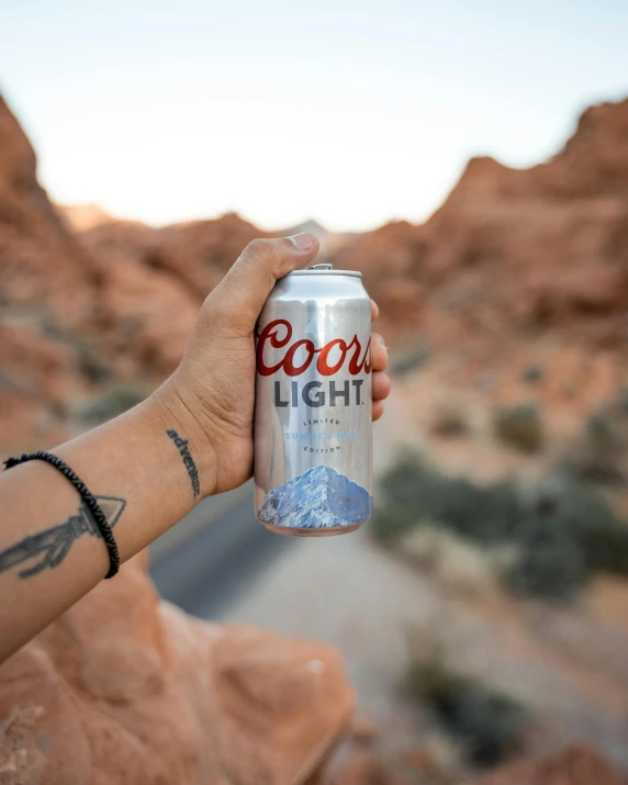 a person holding a can of coors light, by Ryan Pancoast, pexels contest winner, rocky desert, light blues, lgbtq, rock arcs