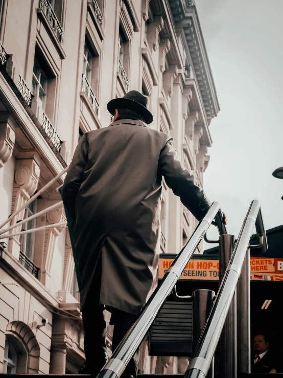 a man walking up an escalator in front of a building, inspired by Louis Stettner, pexels contest winner, wearing a trenchcoat, black stetson and coat, paris hotel style, his back is turned