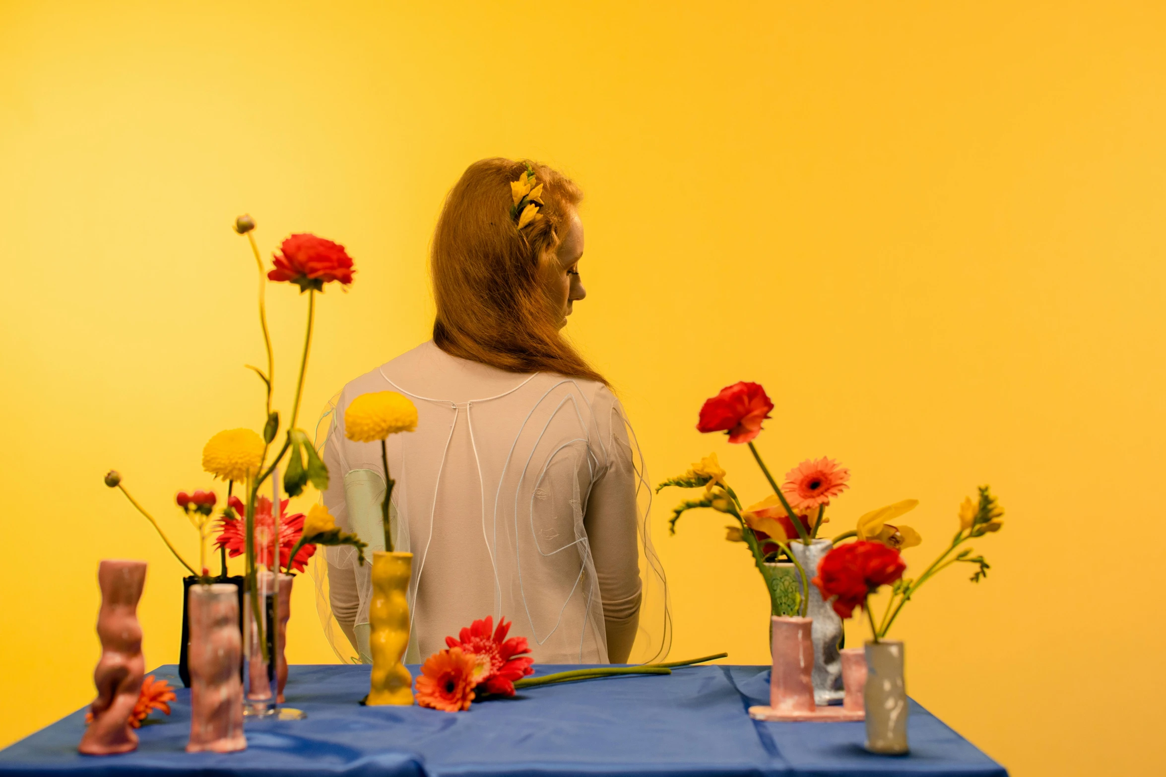 a woman sitting at a table with vases of flowers, an album cover, unsplash, some yellow and blue, ignant, portrait of sanna marin, back towards camera