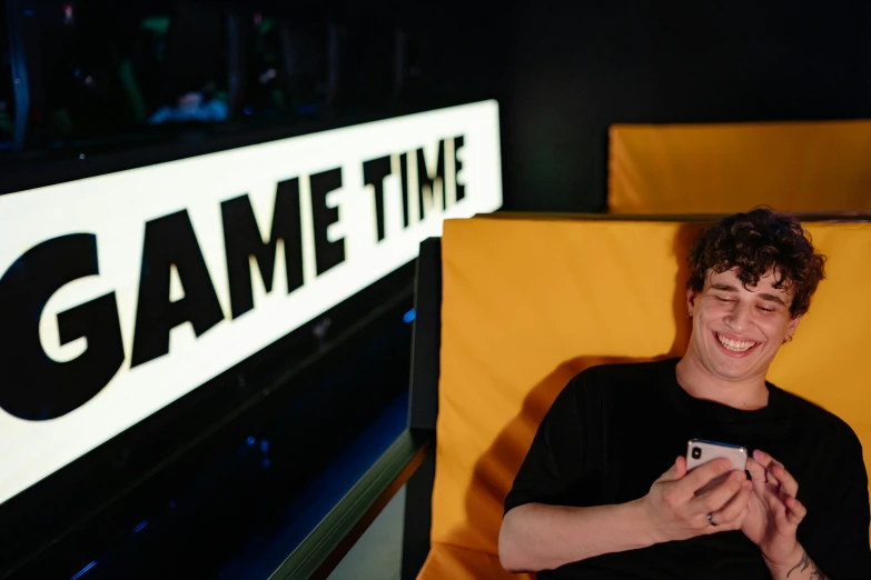 a man sitting in a chair holding a cell phone, an album cover, inspired by Jamie Hewlett, neon letters tripmachine, gaming room, good times photograph, vr game