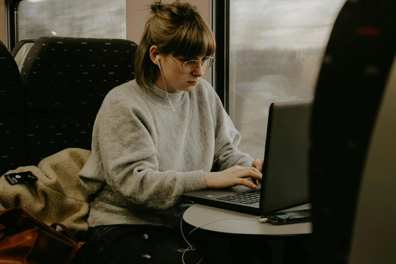 a woman sitting on a train using a laptop computer, a portrait, by Emma Andijewska, trending on pexels, maisie williams, librarian, gaming, wearing a grey hooded sweatshirt