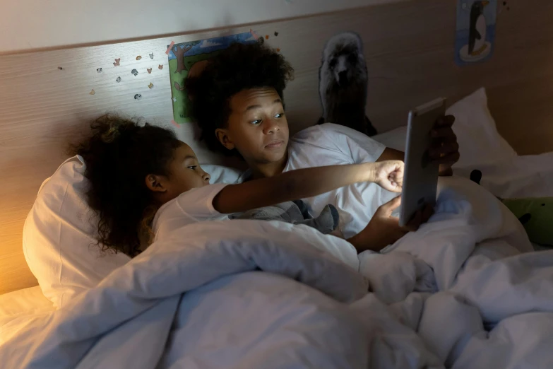 a couple of kids laying in bed next to each other, by Carey Morris, pexels, happening, using a magical tablet, ( ( dark skin ) ), led, bottom angle