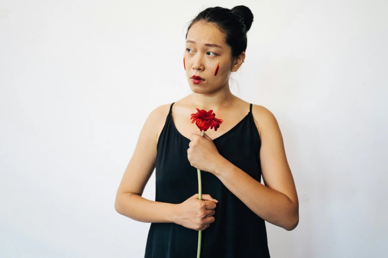 a woman in a black dress holding a red flower, inspired by Ruth Jên, unsplash, antipodeans, prosthetic makeup, young asian woman, with a hurt expression, she is wearing a black tank top