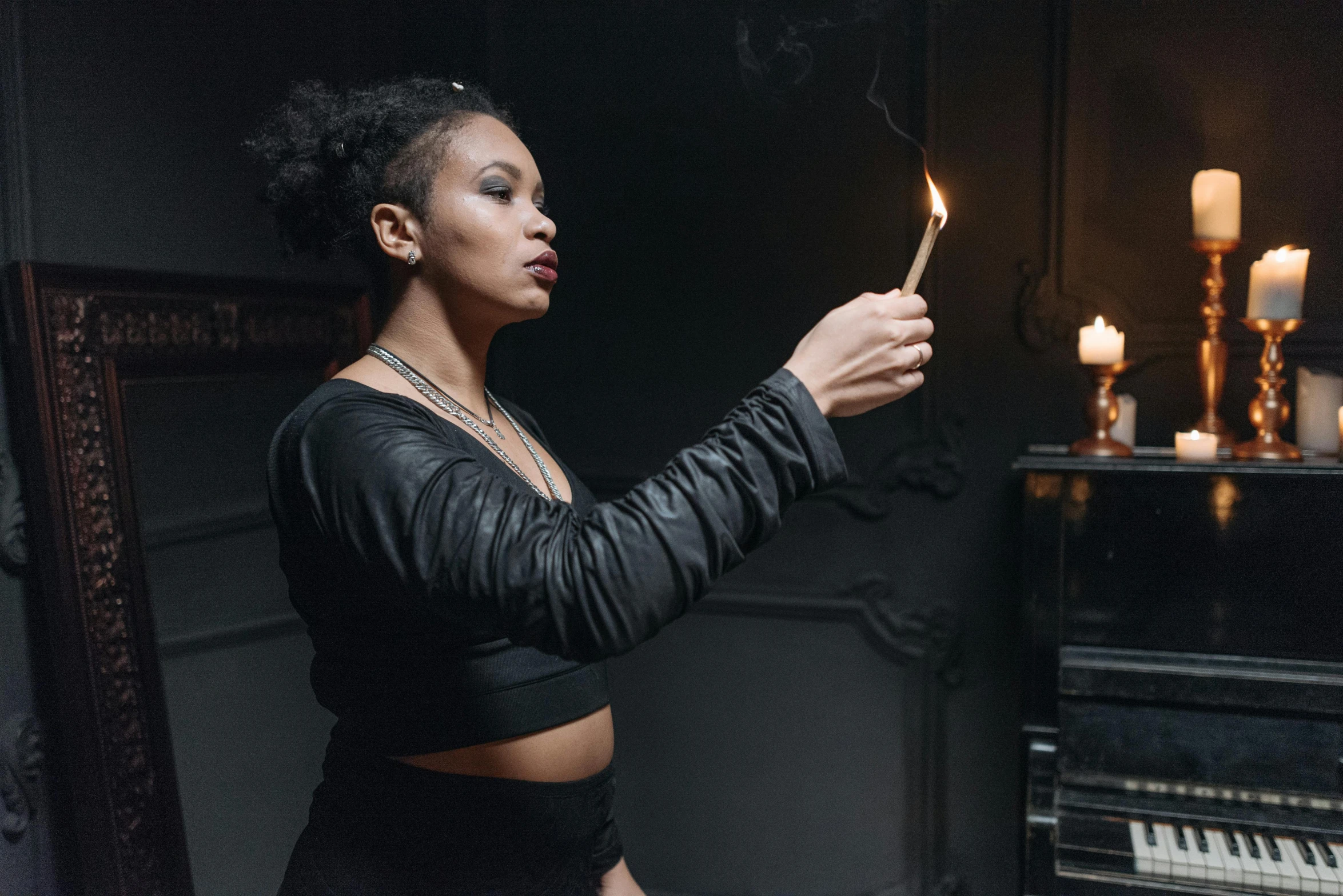 a woman holding a lit candle in front of a piano, inspired by Elsa Bleda, visual art, mage smoking pipe, tessa thompson inspired, gothic clothing, photograph of a techwear woman