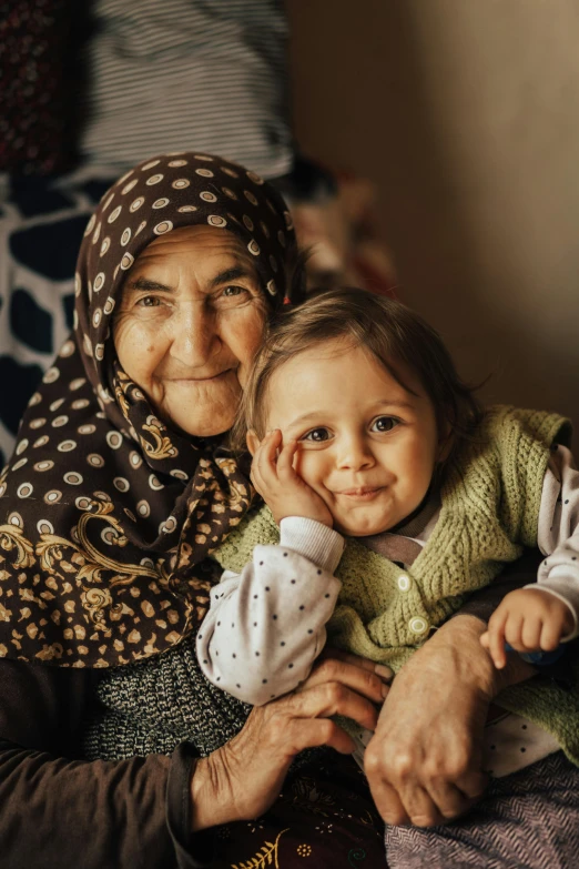 a woman sitting next to a little girl on a bed, by Ibrahim Kodra, pexels contest winner, hurufiyya, elderly woman, lovingly looking at camera, two buddies sitting in a room, turkish and russian