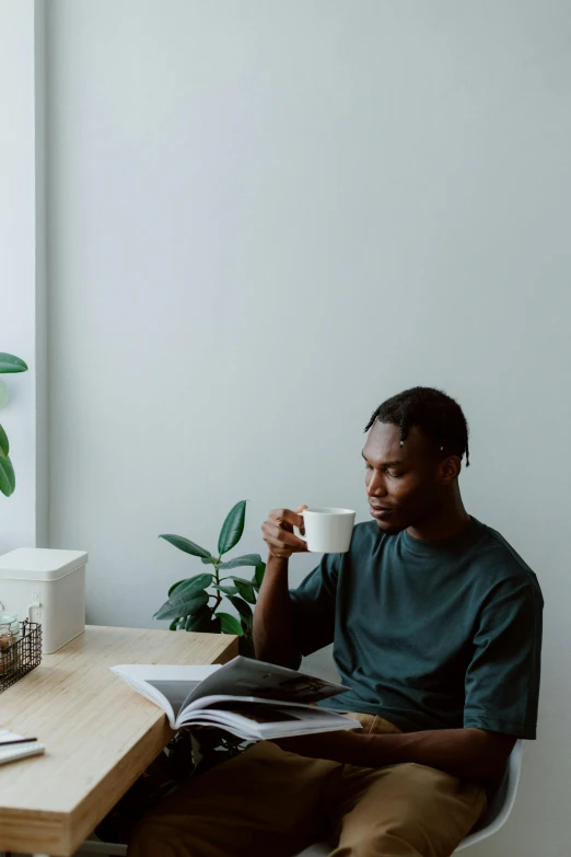 a man sitting at a table with a cup of coffee, dark skinned, next to a plant, slightly minimal, reading