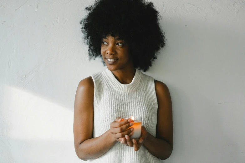 a woman standing in front of a white wall holding a cup, by Carey Morris, pexels contest winner, holding a candle, natural hair, very calm and wholesome, black female