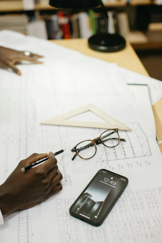 a person sitting at a table with papers and a cell phone, a drawing, inspired by Afewerk Tekle, pexels contest winner, building plans, square rimmed glasses, carefully crafted, carpenter