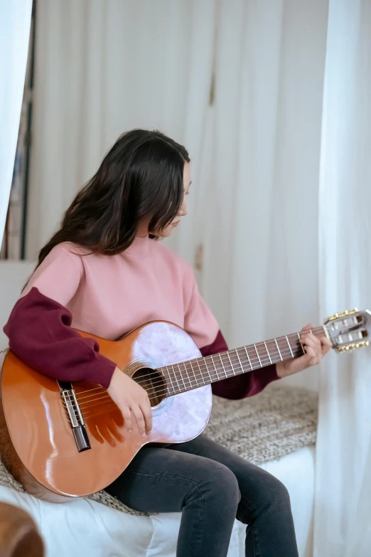 a woman sitting on a bed playing a guitar, pexels contest winner, happening, wearing casual sweater, brown and pink color scheme, mai anh tran, profile pic