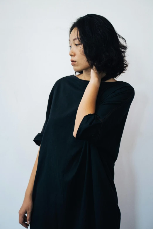 a woman standing in front of a white wall, inspired by Hiromitsu Takahashi, unsplash, black tunic, blackhair, dolman, full product shot