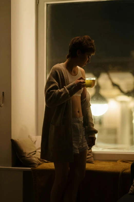 a man standing in front of a window holding a cup, inspired by Elsa Bleda, emma watson in bed, teenage girl, cinestill, lesbians