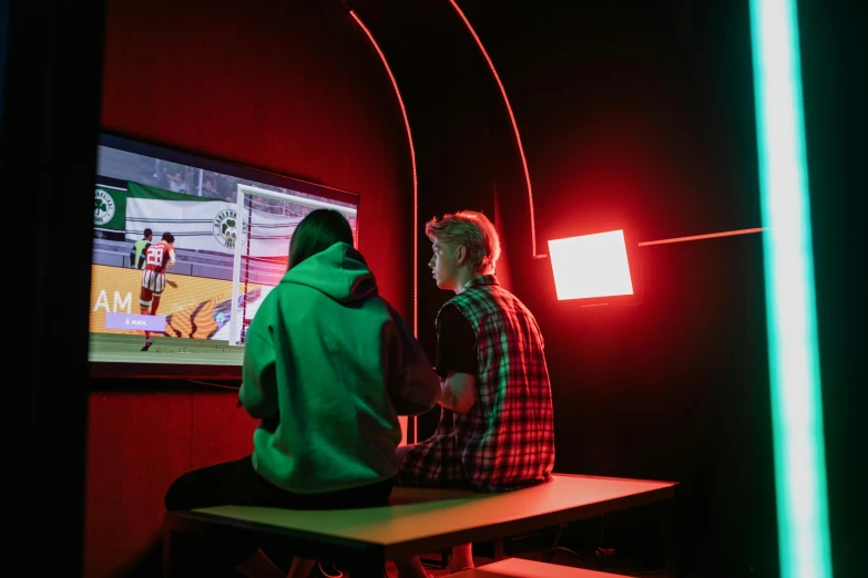 two people sitting at a table in front of a tv, a cartoon, unsplash, interactive art, red and green lighting, sport game, schools, arcade