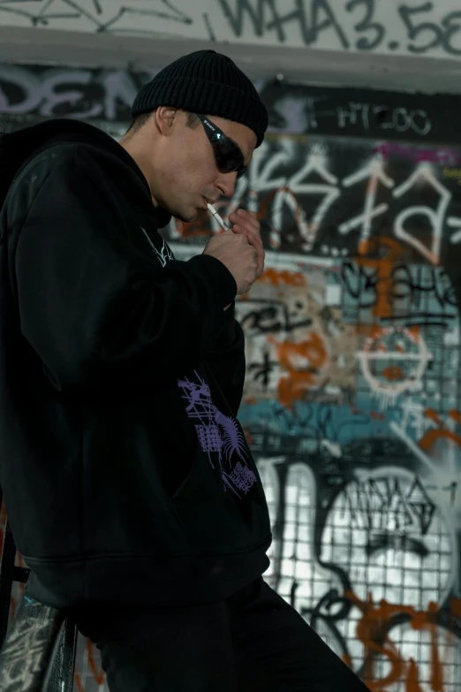 a man smoking a cigarette in front of a graffiti covered wall, wearing a black hoodie, purple drank, large)}], scene