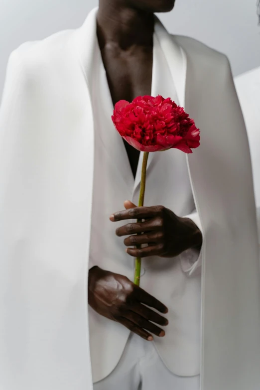 a man in a white suit holding a red flower, inspired by Robert Mapplethorpe, adut akech, long white cape, dark brown skin, nun fashion model