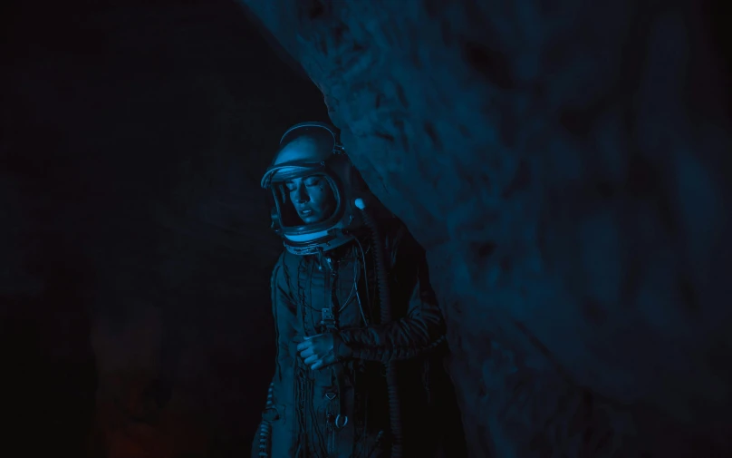 a man in a space suit standing next to a wall, pexels contest winner, in a cave. underexposed, 1 9 6 0 s space girl, navy, dressed as a scavenger