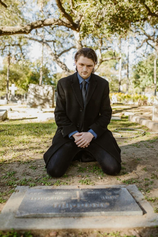 a man sitting on the ground in front of a grave, in a black suit, zach hill, sydney hanson, will graham