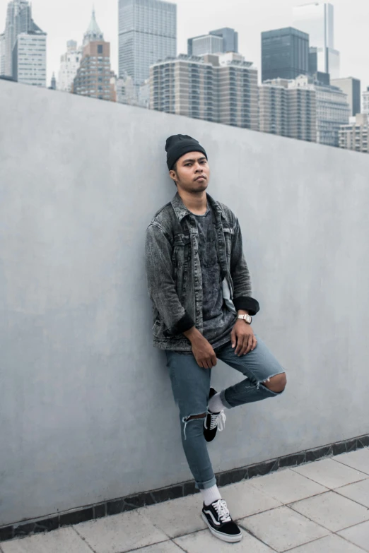 a man leaning against a wall with a city in the background, by Robbie Trevino, wearing jeans and a black hoodie, luis melo, baggy clothing and hat, joel fletcher