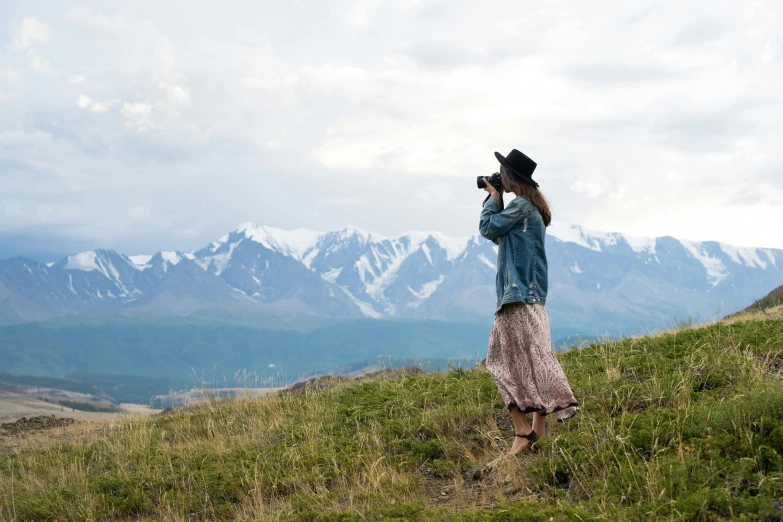 a woman standing on top of a lush green hillside, a picture, inspired by Anka Zhuravleva, unsplash contest winner, icy mountains in the background, holding a camera, profile pose, steppe