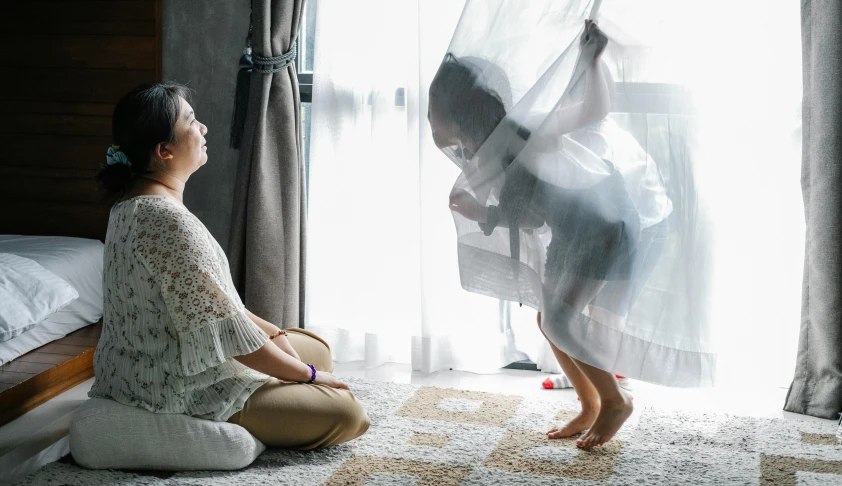 a woman sitting on the floor in front of a window, with a kid, wearing translucent sheet, kids playing, flying shot