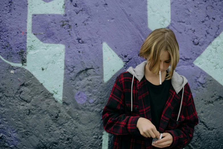 a person standing in front of a wall with graffiti on it, trending on pexels, graffiti, harry potter smoking weed, blond boy, flannel, holding a syringe