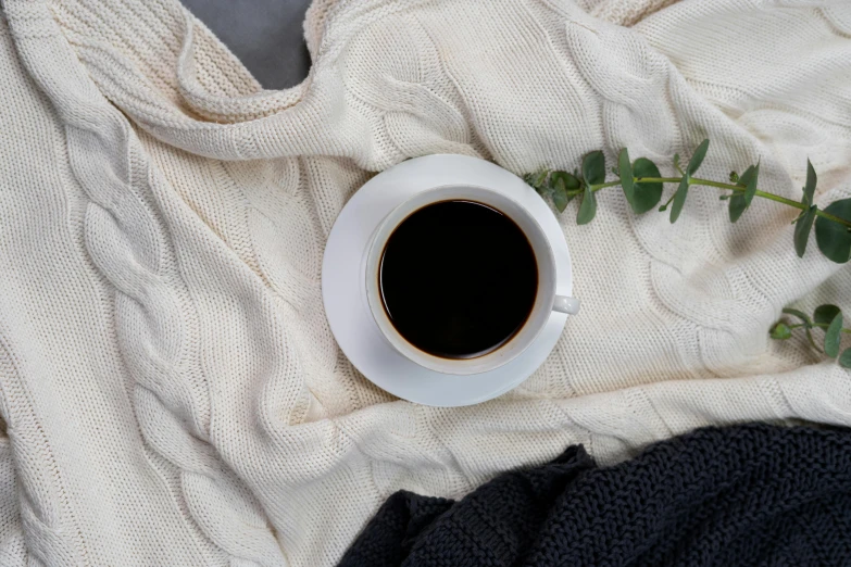 a cup of coffee sitting on top of a blanket, inspired by Ceferí Olivé, trending on pexels, panel of black, wearing a white sweater, sustainable materials, background image