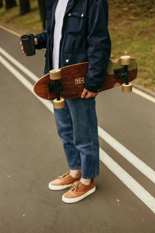 a man standing on the side of a road holding a skateboard, brown pants, holding a drink, top down shot, duck shoes