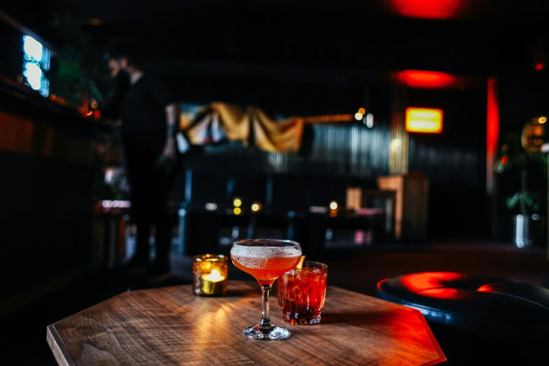 a couple of glasses sitting on top of a wooden table, unsplash, speakeasy bar background, an island made of red caviar, in chippendale sydney, female death holding a cocktail