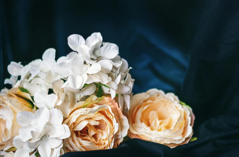 a bouquet of flowers sitting on top of a blue cloth, by Sylvia Wishart, unsplash, romanticism, gold and white robes, hydrangea, pale orange colors, with a black dark background