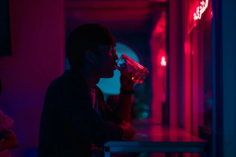 a man sitting at a table drinking from a glass, inspired by Liam Wong, pexels contest winner, serial art, pink and blue lighting, neon standup bar, atmospheric red lighting, high school