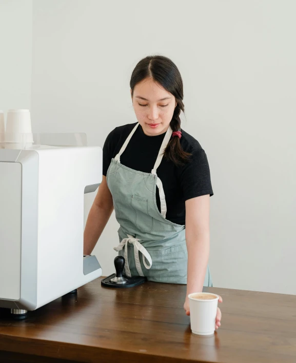 a woman standing in front of a coffee machine, by Jessie Algie, trending on unsplash, private press, white waist apron and undershirt, on a table, plain background, background image